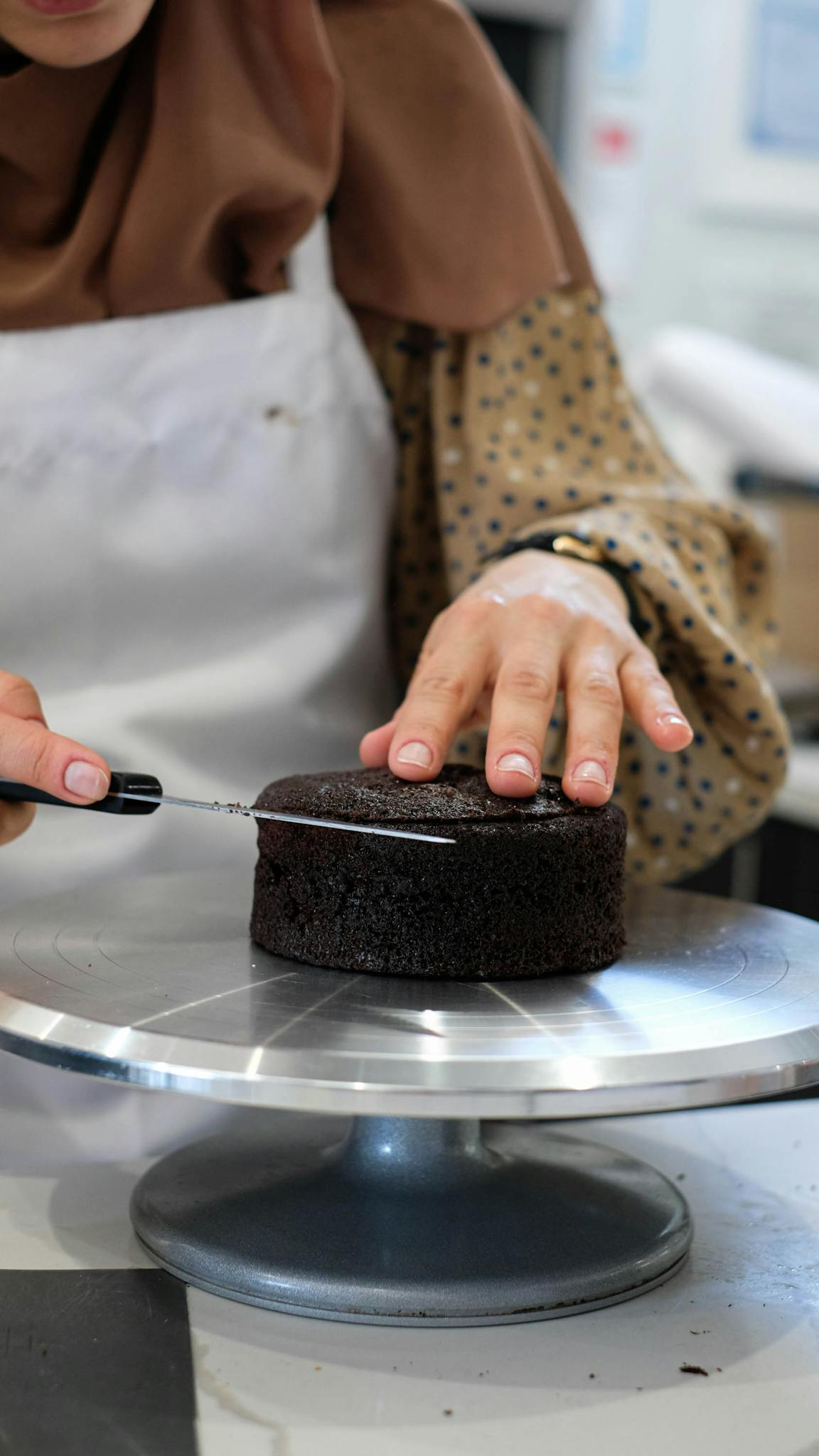 A woman precisely slicing a chocolate cake on a turntable indoors, showcasing skilled baking techniques.